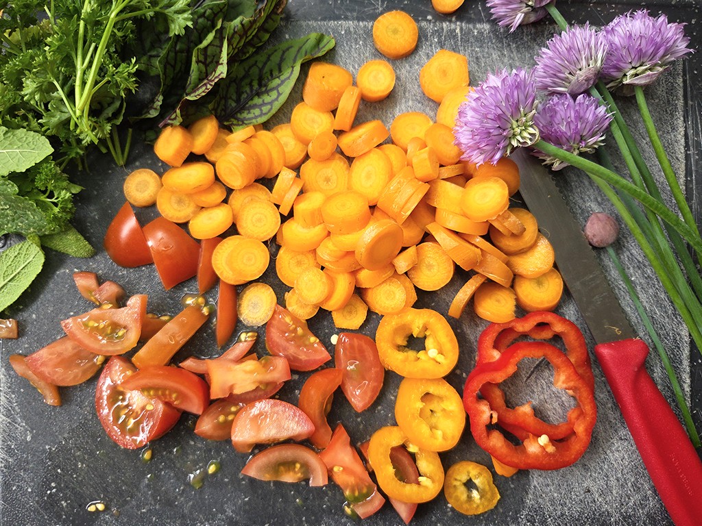 Chopped orange carrot coins, red bell pepper rings, and tomato wedges spread on a dark cutting board with greens on the side and purple allium flowers nearby.