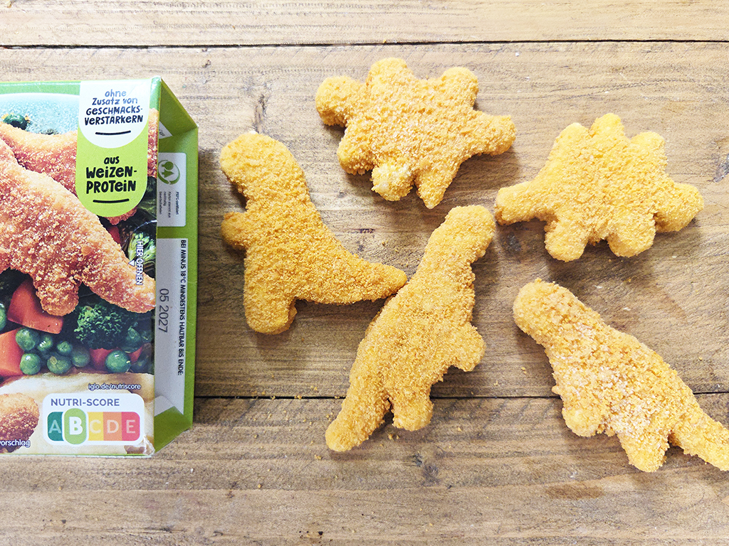 Dinosaur-shaped breaded chicken nuggets spread on a wooden table beside a snack box.