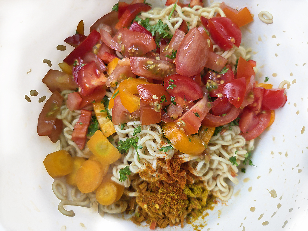 Noodles topped with chopped tomatoes, bell peppers, and carrots, garnished with parsley and a brown spice crumb.