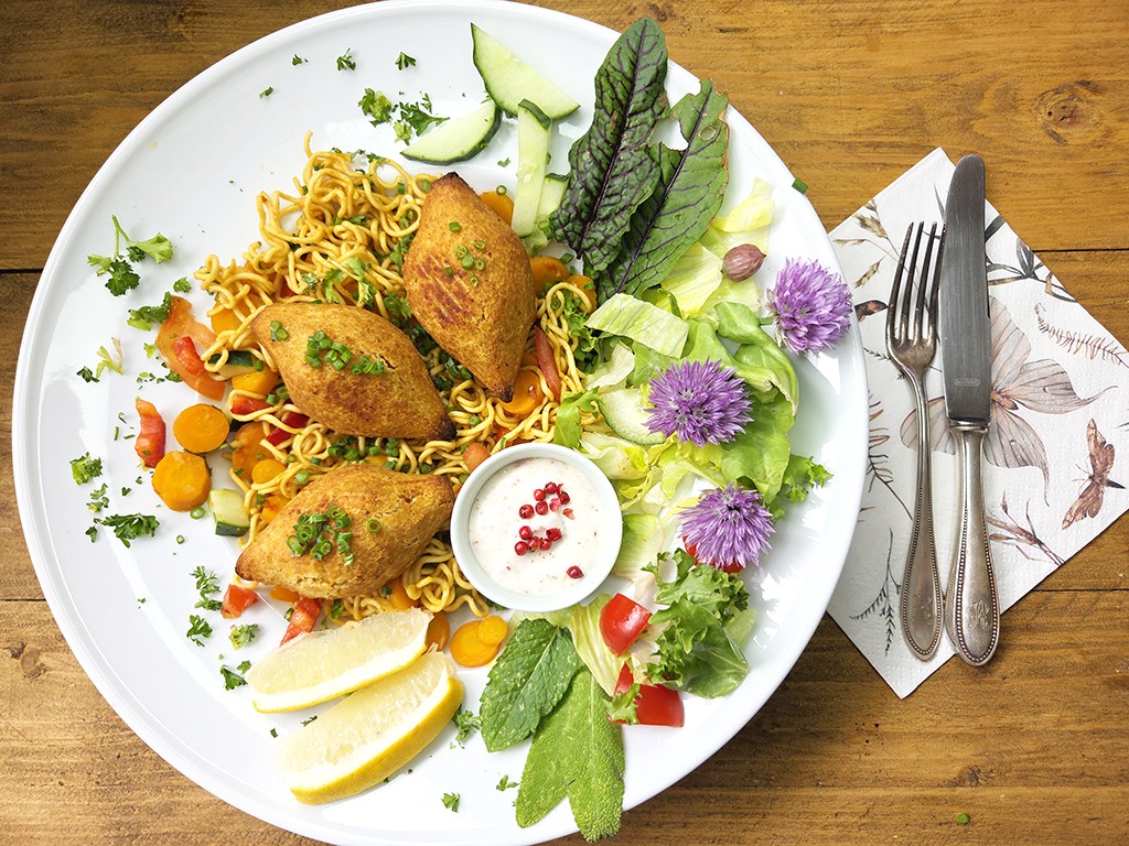 Plate of fried falafel on noodle salad with lemon wedges, mixed greens, cucumber, carrot rounds, and purple flowers; dipping sauce in center.