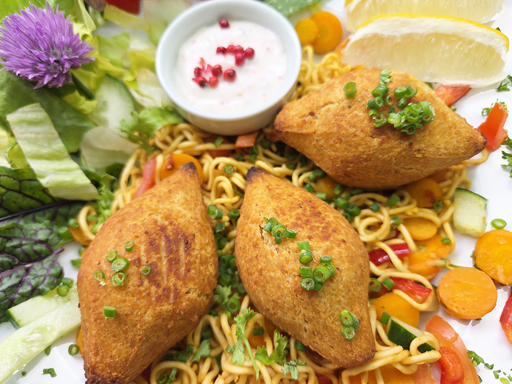 Three golden fried samosas resting on a bed of noodles with diced vegetables, garnished with chopped chives and herbs, plus dipping sauce and lemon wedge.