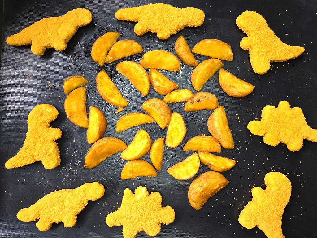 Assorted dinosaur-shaped crackers surround a pile of oven-baked potato wedges on a dark baking tray.
