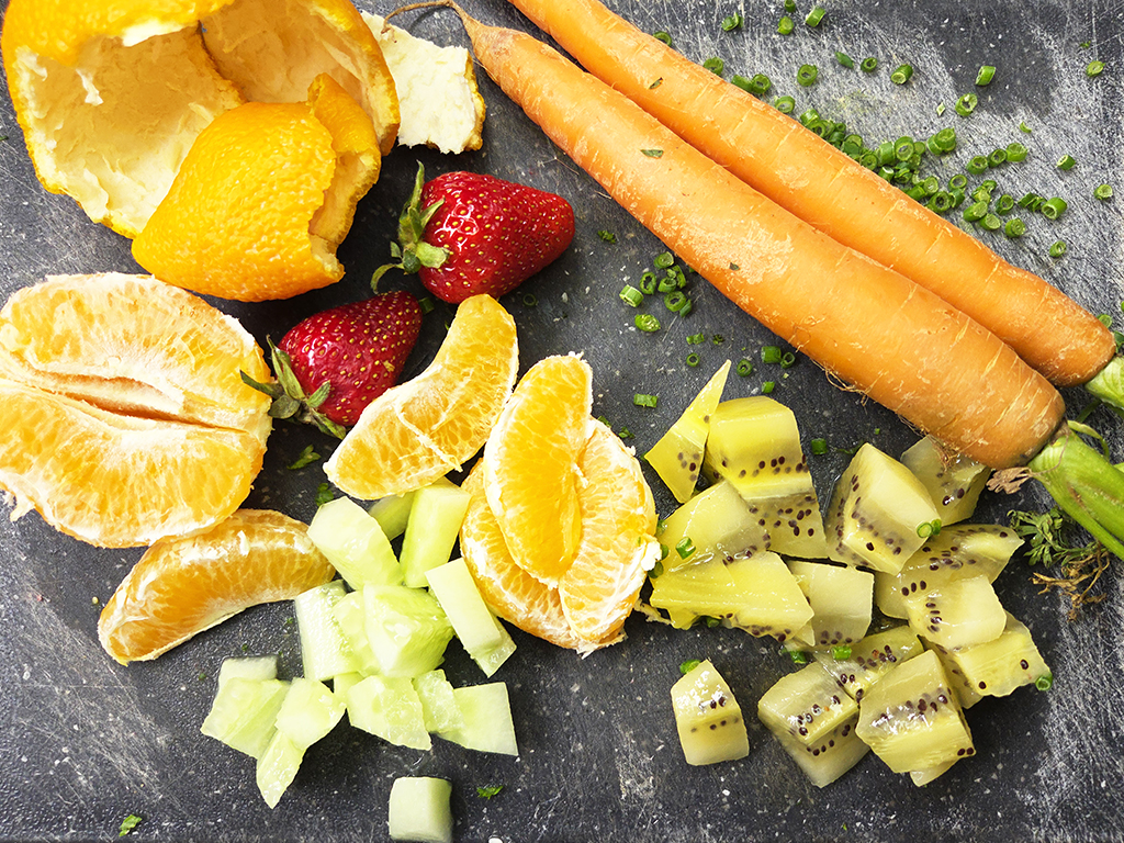 Colorful fruit platter with orange segments, strawberries, kiwi chunks, melon cubes, and whole carrots on a dark surface.