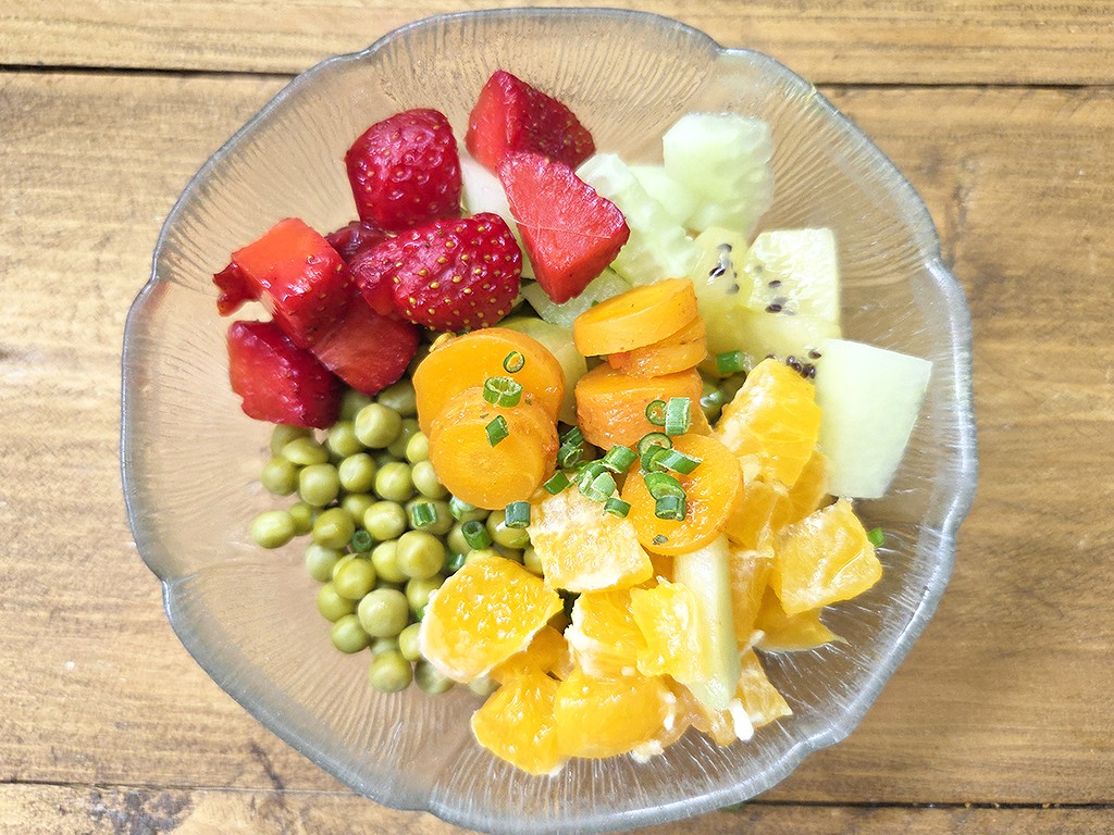 Fruit salad in a clear glass bowl on a wooden surface, featuring strawberries, orange slices, pineapple, melon, peas, and chives as garnish.