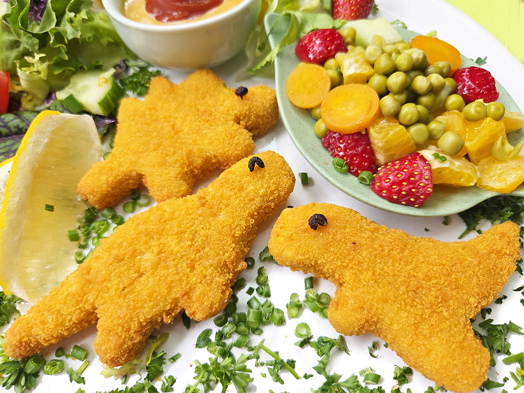 Two golden, breaded fish-shaped nuggets on a plate with a lemon wedge, green herbs, and a side of mixed fruit salad and greens.