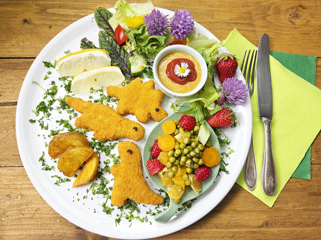 Top-down view of a large plate with lemon wedges, mixed greens, orange breaded chicken shapes, potato wedges, a side of veggie salad with peas and corn, strawberries, and a small dipping sauce, with colorful napkins and cutlery nearby.