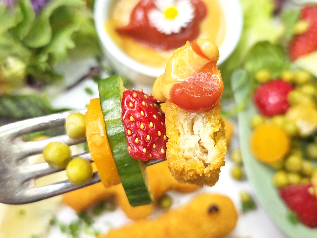 Fork with a skewer of cucumber, orange slice, and strawberry, plus a crispy fried bite with sauce on top; colorful plate in background.
