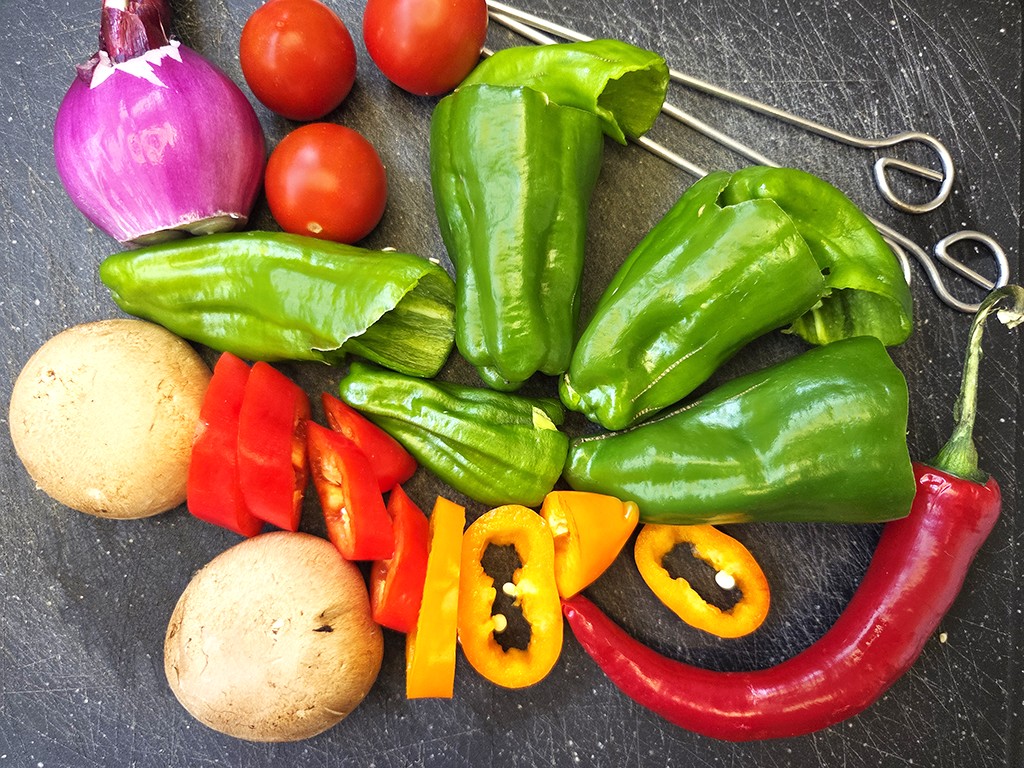 Wheaty | Bio BBQ Green Piece Seitan | Herzschatz Assorted fresh vegetables on a dark cutting board: red onion, cherry tomatoes, green bell peppers, sliced red and yellow peppers, a red chili, round mushrooms, and kitchen scissors in the background.