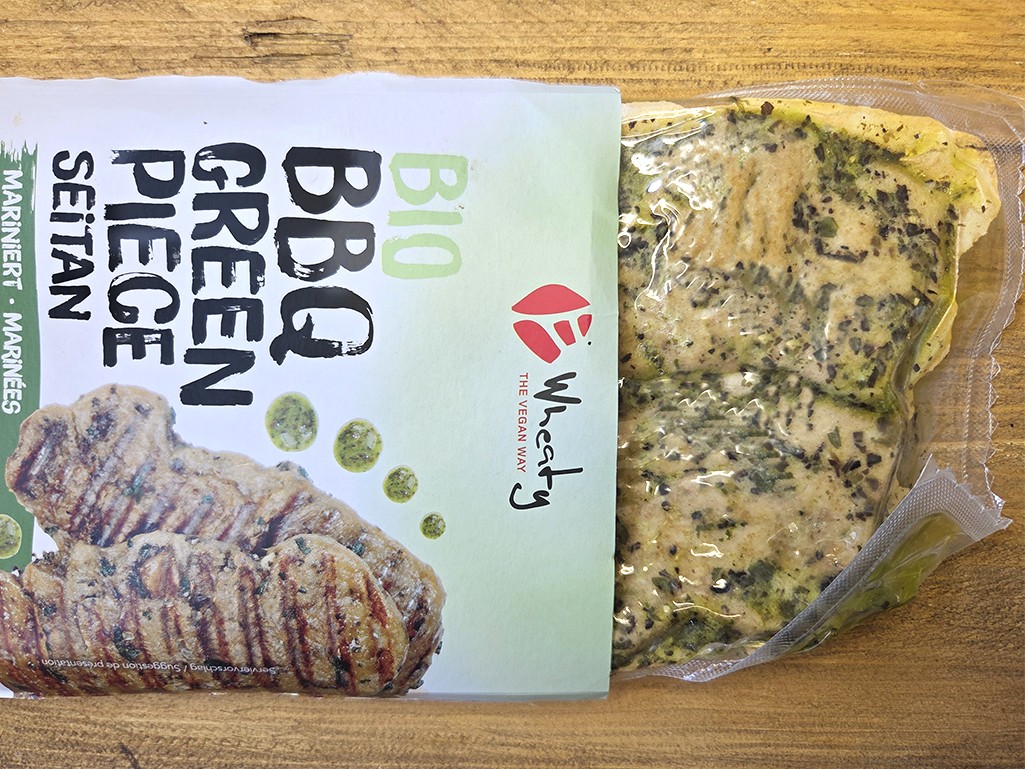 Wheaty | Bio BBQ Green Piece Seitan | Herzschatz Open package of vegan veggie burger patties on a wooden surface; herb-seasoned patties visible in vacuum-sealed bag.