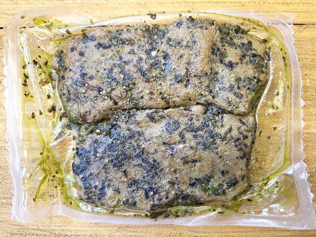 Wheaty | Bio BBQ Green Piece Seitan | Herzschatz Two herb-seasoned fish fillets in a sealed plastic pouch with oil on a wooden surface.