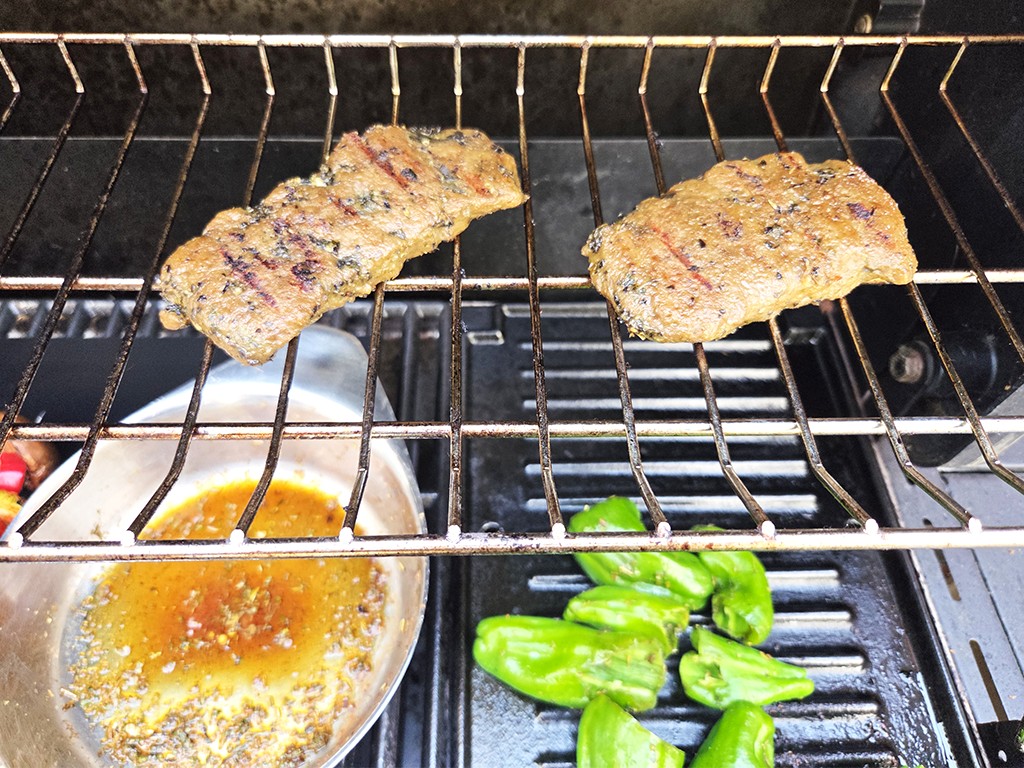 Wheaty | Bio BBQ Green Piece Seitan | Herzschatz Two grilled meat pieces on a rack above a pan of sauce, with green peppers on the lower grill rack nearby.