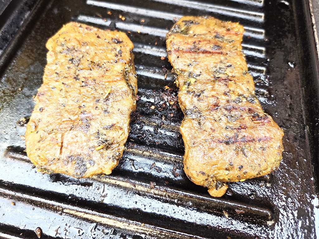 Wheaty | Bio BBQ Green Piece Seitan | Herzschatz Two herb-seasoned burger patties cooking on a grill pan with visible sear marks and oil around them.