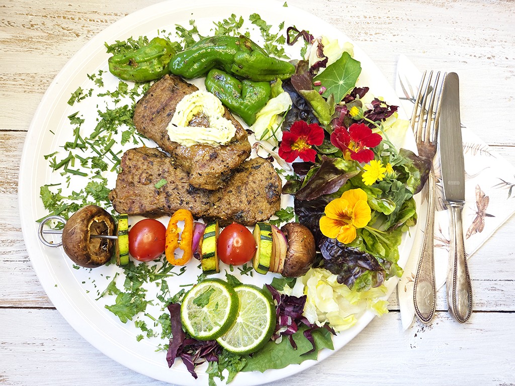 Wheaty | Bio BBQ Green Piece Seitan | Herzschatz Plate with grilled meat patties, veggie kebabs, and a fresh greens salad with lime wedges and edible flowers.