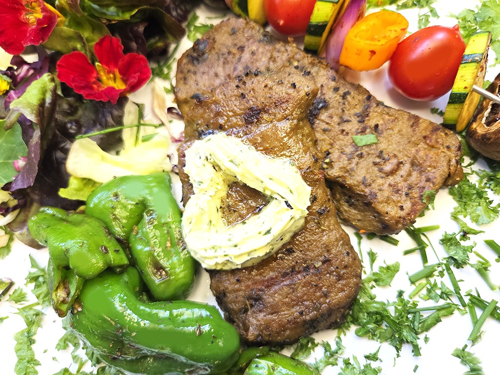 Wheaty | Bio BBQ Green Piece Seitan | Herzschatz Beef steak slices topped with herb butter, served with sautéed green peppers, a veggie skewer, and a leafy salad on a garnished plate.