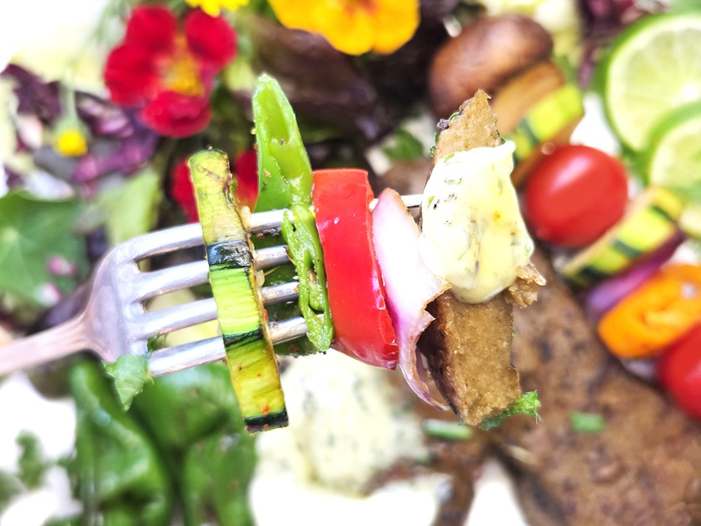 Wheaty | Bio BBQ Green Piece Seitan | Herzschatz Fork with a skewer of grilled vegetables (zucchini, green pepper, tomato, onion) and a bread bite with herb butter against a colorful background.