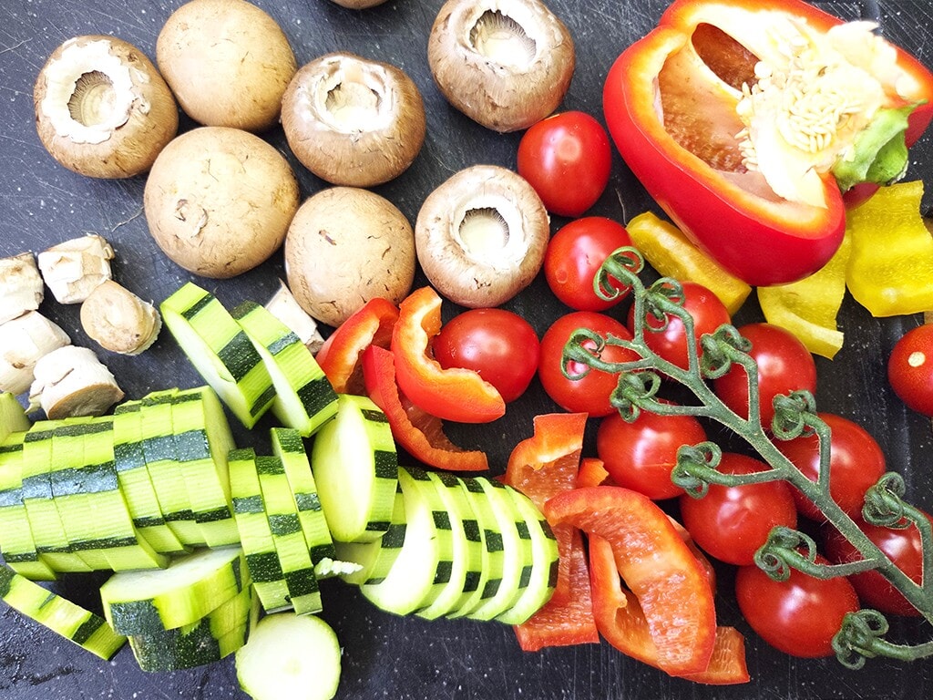 Revo | El Pollo | Herzschatz Assorted chopped vegetables on a dark cutting board: mushrooms, zucchini, cherry tomatoes, red and yellow peppers ready for cooking.