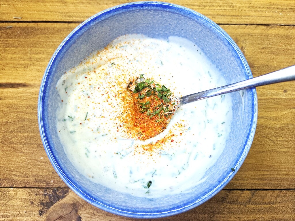 Revo | El Pollo | Herzschatz Blue bowl of creamy herb yogurt sauce with paprika and chopped herbs, on a wooden table with a spoon resting inside.