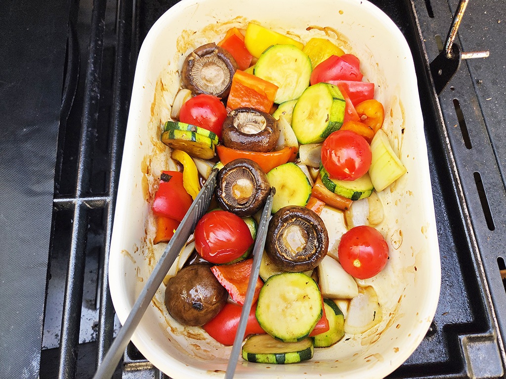 Revo | El Pollo | Herzschatz White baking dish of roasted vegetables on a grill: tomatoes, sliced zucchini, bell peppers, mushrooms and onions with tongs resting inside the dish
