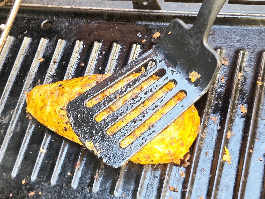 Revo | El Pollo | Herzschatz Orange-tinted chicken breast on a hot grill being pressed by a slotted spatula to sear evenly.