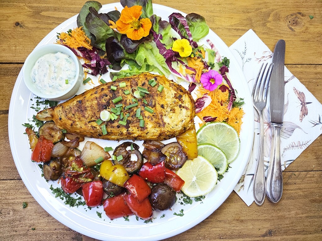 Revo | El Pollo | Herzschatz Herb-roasted chicken leg with peppers and onions, mixed greens with edible flowers, lime wedges, and a side of sauce on a white plate.