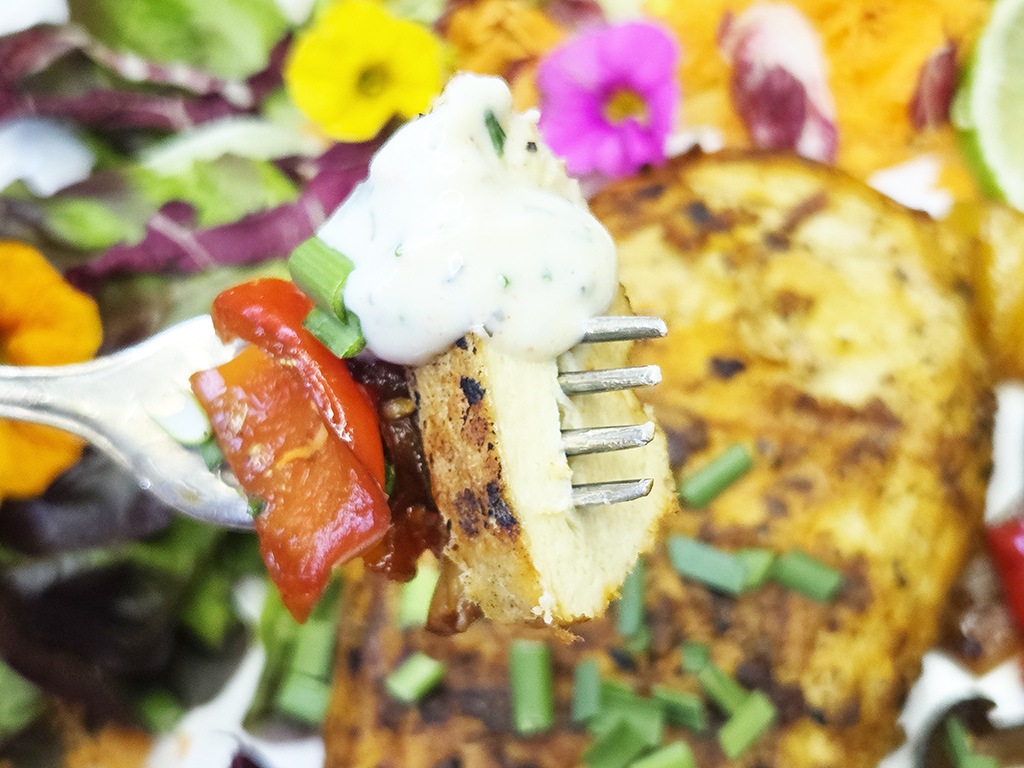 Revo | El Pollo | Herzschatz Close-up of a fork holding a toasted bread slice topped with tomato, herb cream sauce, and chives, with a colorful dish in the background.