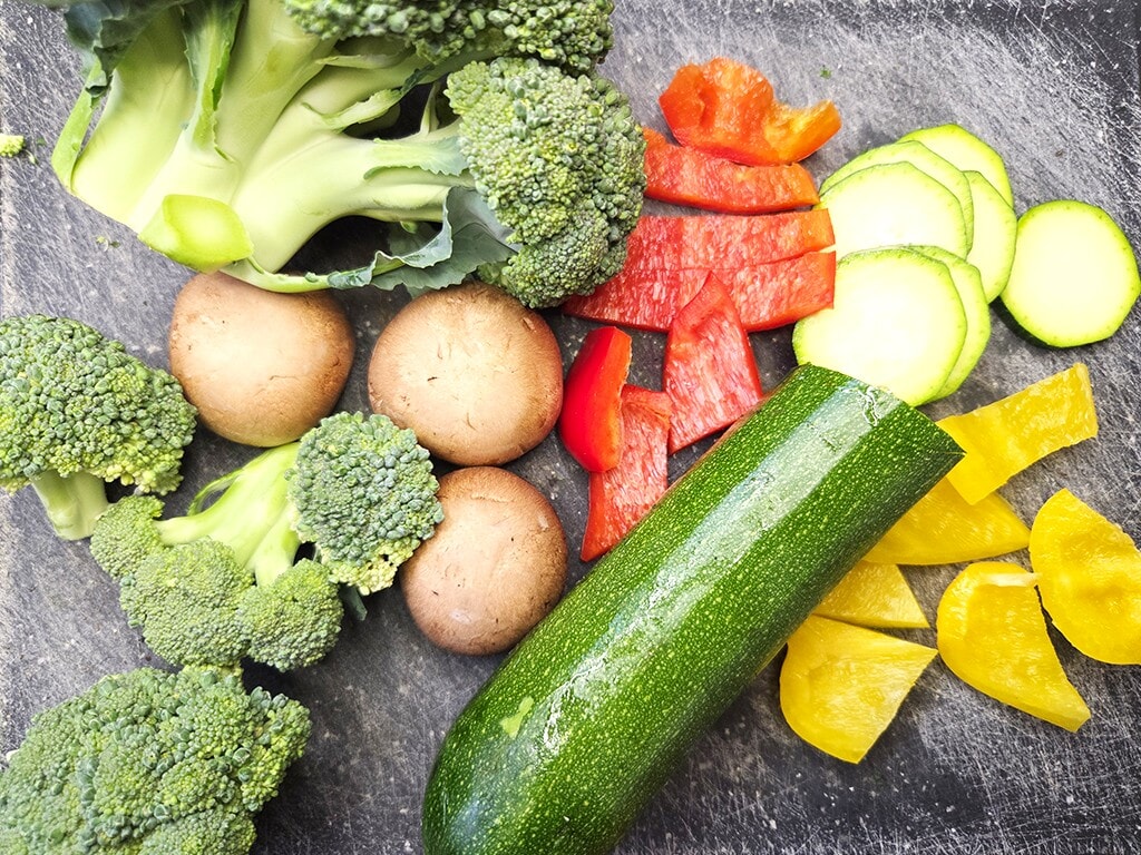 Hengstenberg | Bulgur Salat mit Tomate und Karotte | Herzschatz Assorted fresh vegetables on a dark cutting board: broccoli florets, mushrooms, sliced red and yellow peppers, zucchini, and cucumber rounds.