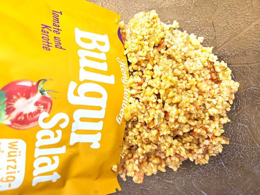 Hengstenberg | Bulgur Salat mit Tomate und Karotte | Herzschatz Bright yellow package labeled Bulgur Salat beside a mound of prepared bulgur salad on a dark textured surface