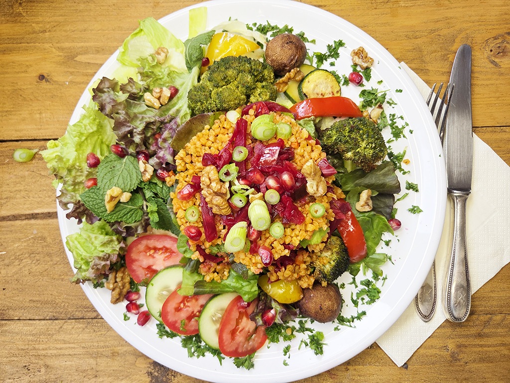 Hengstenberg | Bulgur Salat mit Tomate und Karotte | Herzschatz Plate of colorful mixed salad with grain center, broccoli, peppers, greens, cucumber, tomatoes, walnuts, and pomegranate seeds on a wooden table.