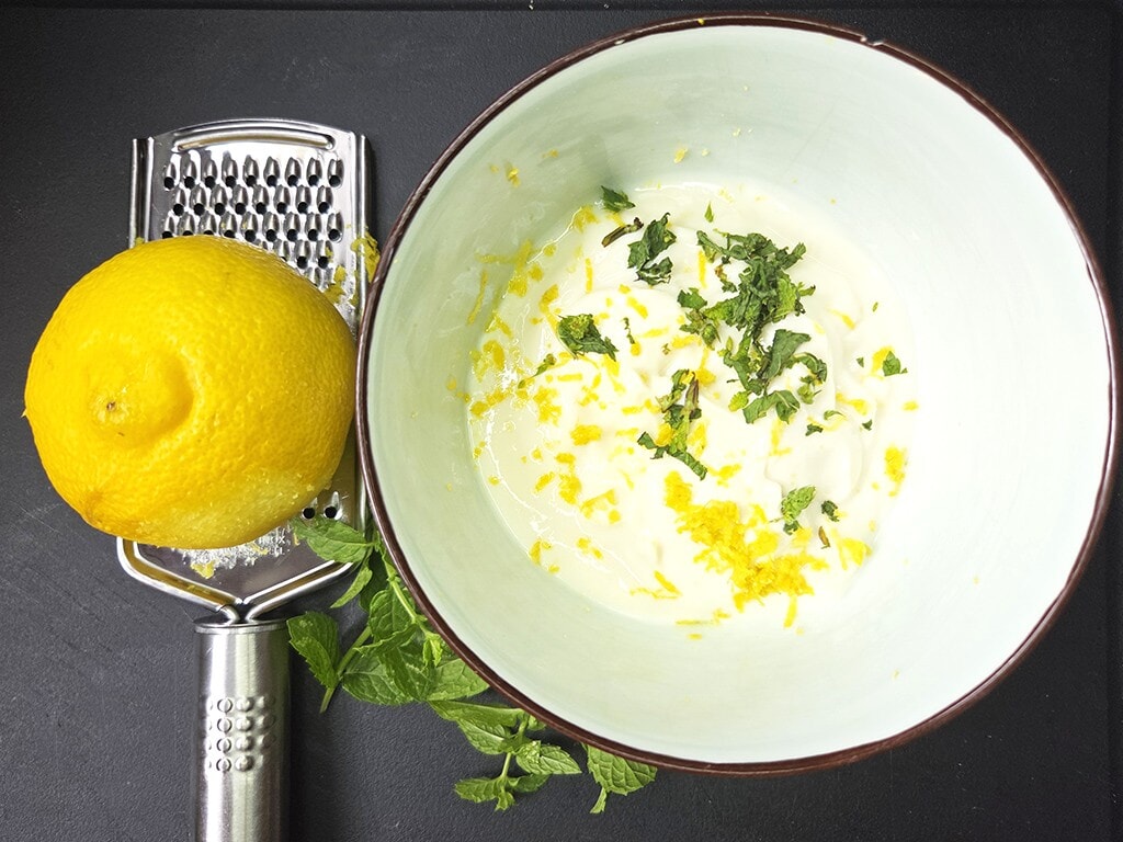 Fresh lemon and a grater beside a bowl of yogurt topped with lemon zest and chopped mint.