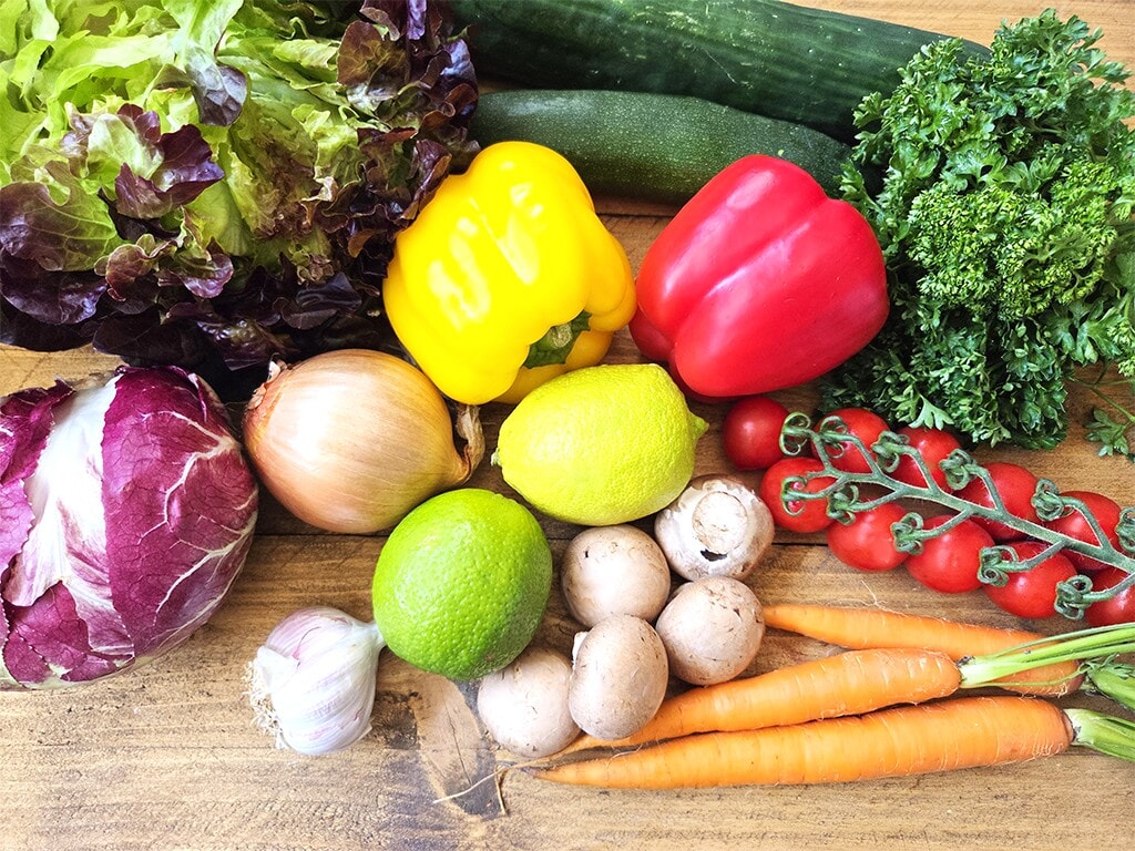 Revo | El Pollo | Herzschatz Colorful display of fresh vegetables and citrus on a wooden surface, including peppers, tomatoes, carrots, greens, mushrooms, onion, garlic, lemon, and lime.