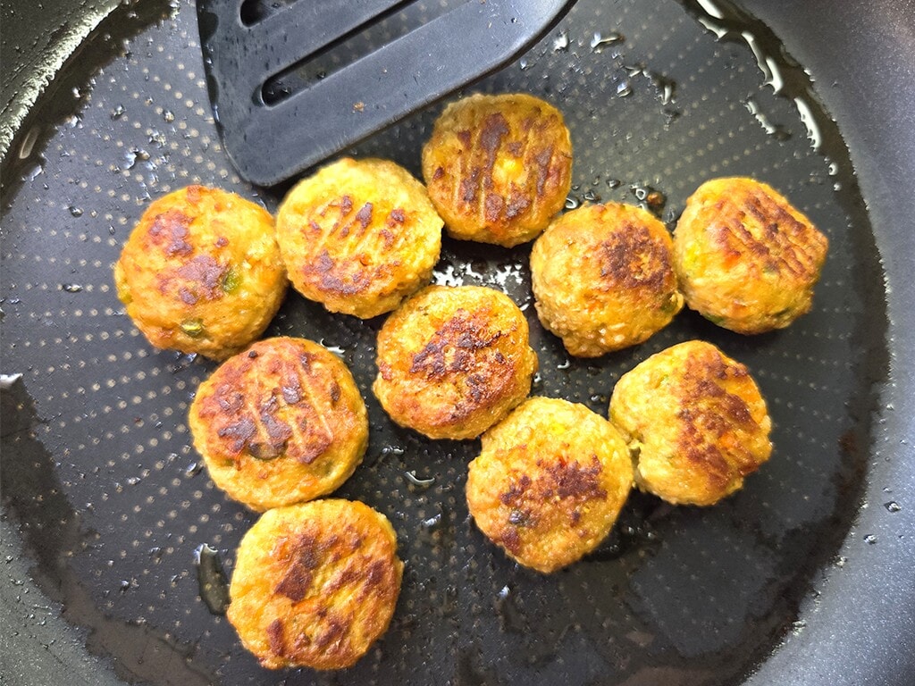 Golden brown round veggie patties frying in a nonstick pan with a spatula nearby.