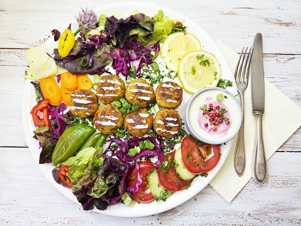 Colorful plate of falafel balls with mixed greens, vegetables, lemon slices, and a small bowl of yogurt-dill sauce on the side