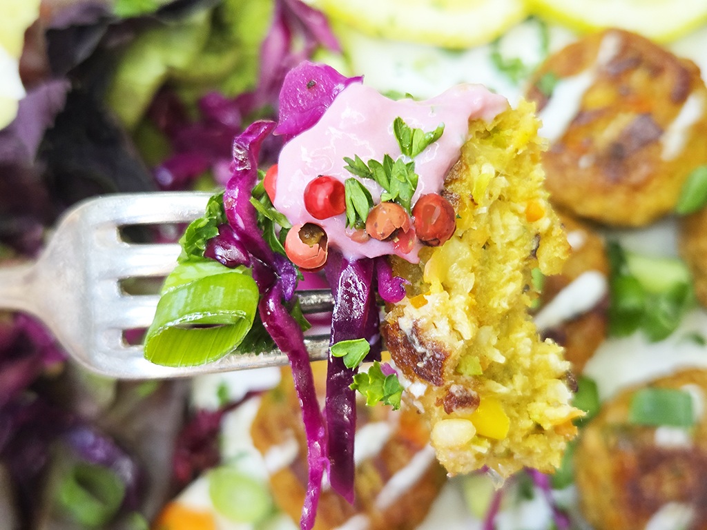 Fork holding a colorful veggie patty bite with pink sauce, greens, purple cabbage, and red peppercorns.