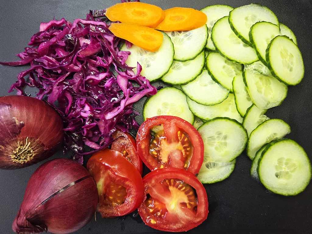 Sliced cucumbers arranged along the right, with red onion halves, shredded purple cabbage, carrot slices, and tomato rounds on a dark surface.