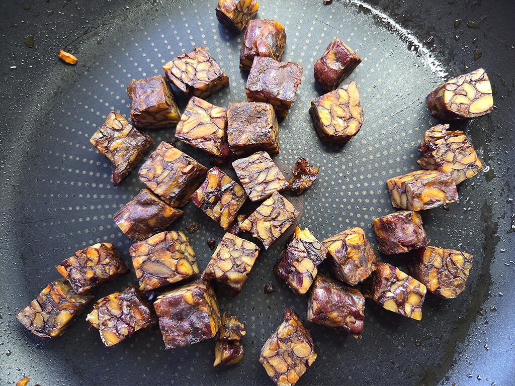 Small squares of peanut brittle with visible peanut pieces, cooling in a dark pan.
