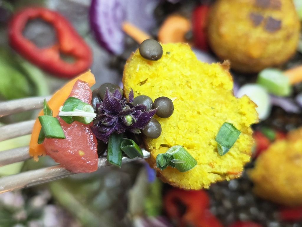 SOTO | Falafel Traditionell | Herzschatz Close-up of a yellow cornmeal bite on a fork, topped with tomato, chopped green onions, a purple flower, capers, and greens.
