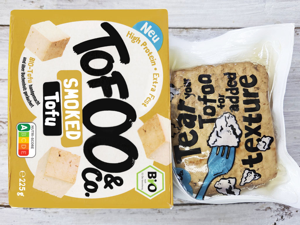 Tofoo & Co | Smoked Tofu | Herzschatz Bright yellow box of smoked tofu snacks beside a sealed bag of cookies on a wooden surface, both packaged foods with bold graphics and text.