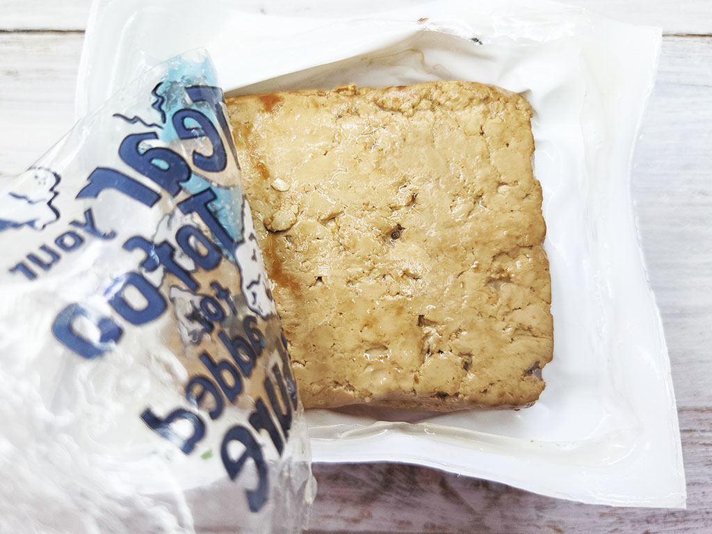 Tofoo & Co | Smoked Tofu | Herzschatz A square, golden-brown cookie bar on a white tray, partially unwrapped from a blue-and-white plastic bag coming in from the left.