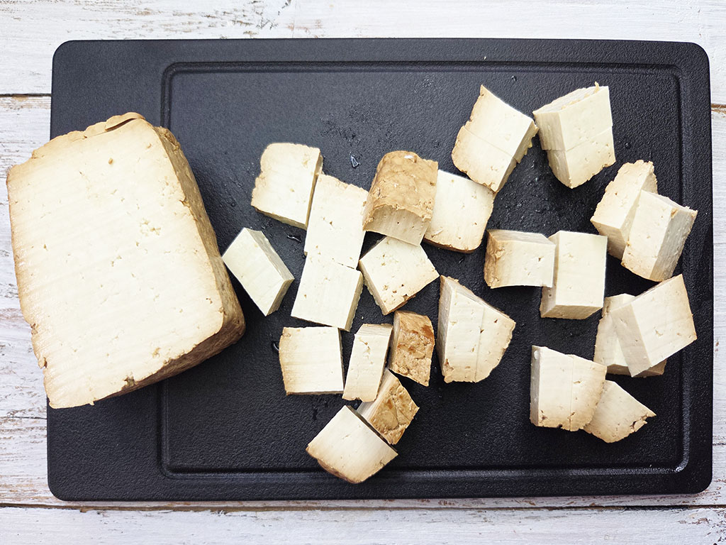 Tofoo & Co | Smoked Tofu | Herzschatz Large block of tofu on a black cutting board with numerous cubed pieces scattered around for cooking prep.