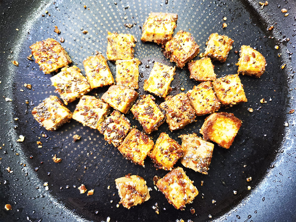 Tofoo & Co | Smoked Tofu | Herzschatz Cubes of crispy browned tofu with sesame seeds frying in a black skillet.