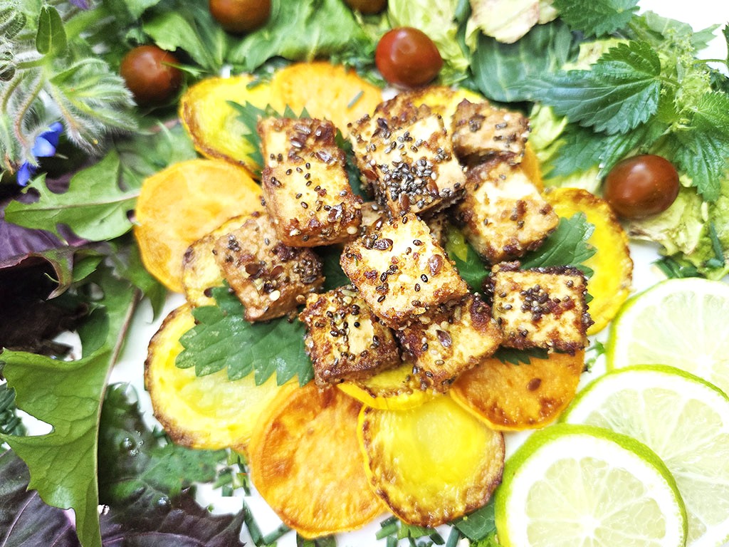 Tofoo & Co | Smoked Tofu | Herzschatz Crispy sesame-coated tofu cubes on a bed of roasted sweet potato rounds, with mixed greens, cherry tomatoes and lime wedges.
