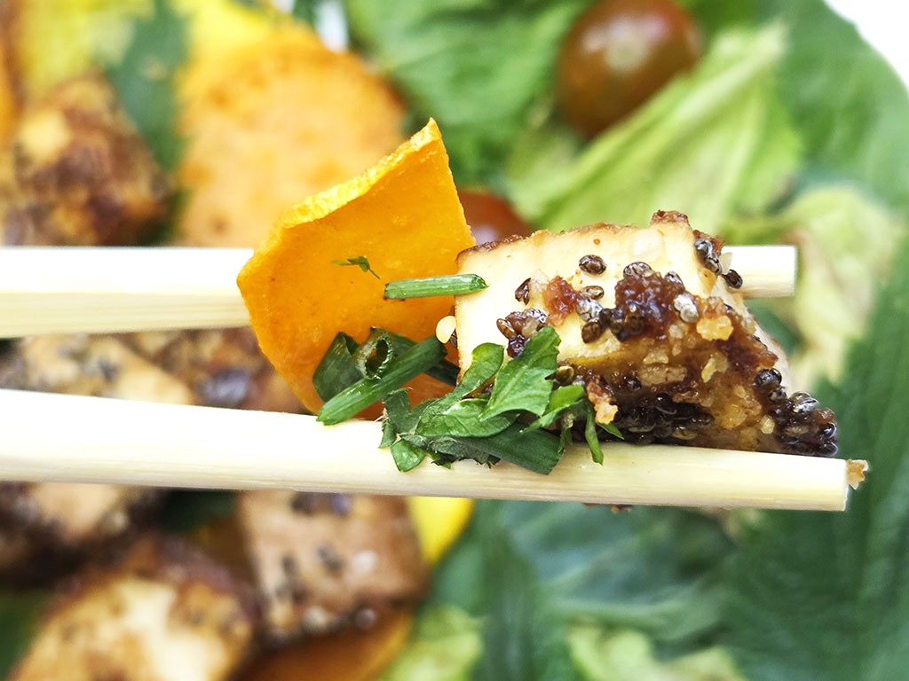 Tofoo & Co | Smoked Tofu | Herzschatz Close-up of a bite-sized fried tofu nugget with sesame seeds and herbs held on chopsticks above a green salad and an orange chip.