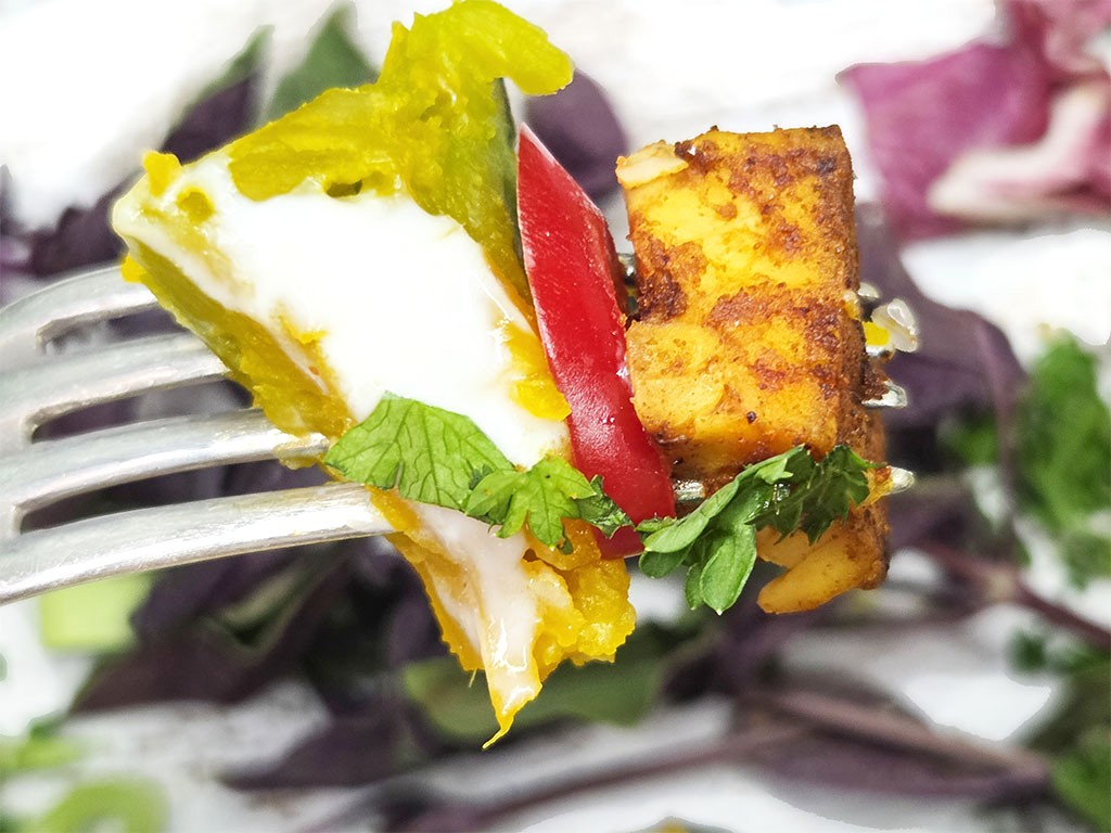 Fork holding a bite of breakfast: fried egg, potato hash cubes, red pepper slice, and herbs on a leafy background