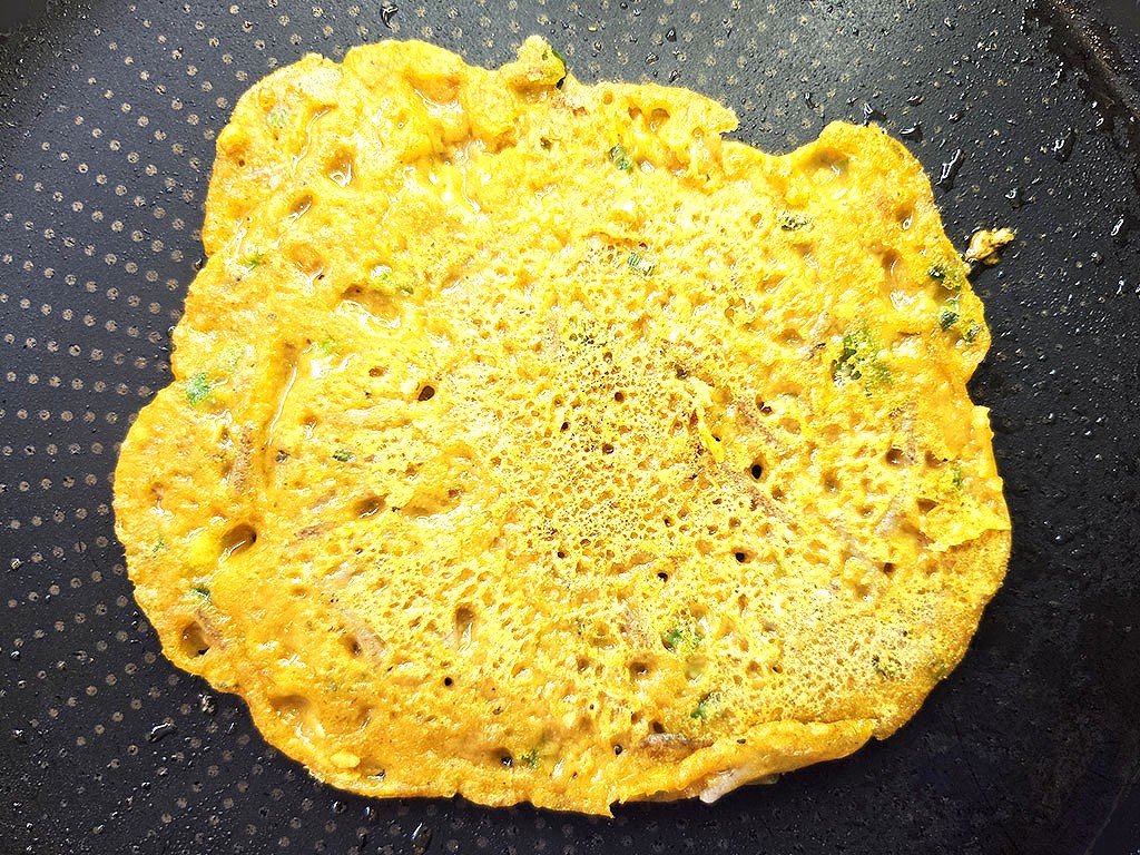 Planted. | Filet-Würfel a la mexicana | Herzschatz Golden-yellow savory pancake (besan chilla) frying in a black skillet with green herbs.