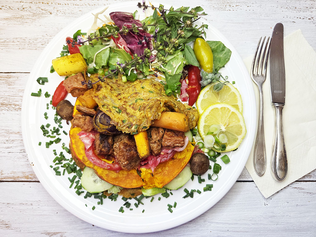 Planted. | Filet-Würfel a la mexicana | Herzschatz White plate featuring a colorful dish: mixed greens and herbs, roasted squash, peppers, mushrooms, and meat, with lemon slices on the side and cutlery nearby.