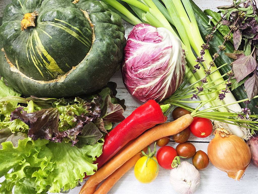 Assorted fresh vegetables on a white wooden table: pumpkin, radicchio, peppers, carrots, onions, garlic, and leafy greens.
