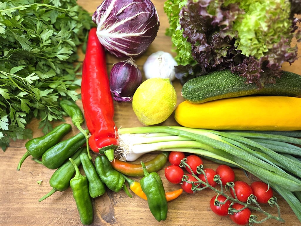 Assorted fresh vegetables on a wooden surface: peppers, tomatoes, leafy greens, cabbage, zucchini, and garlic.