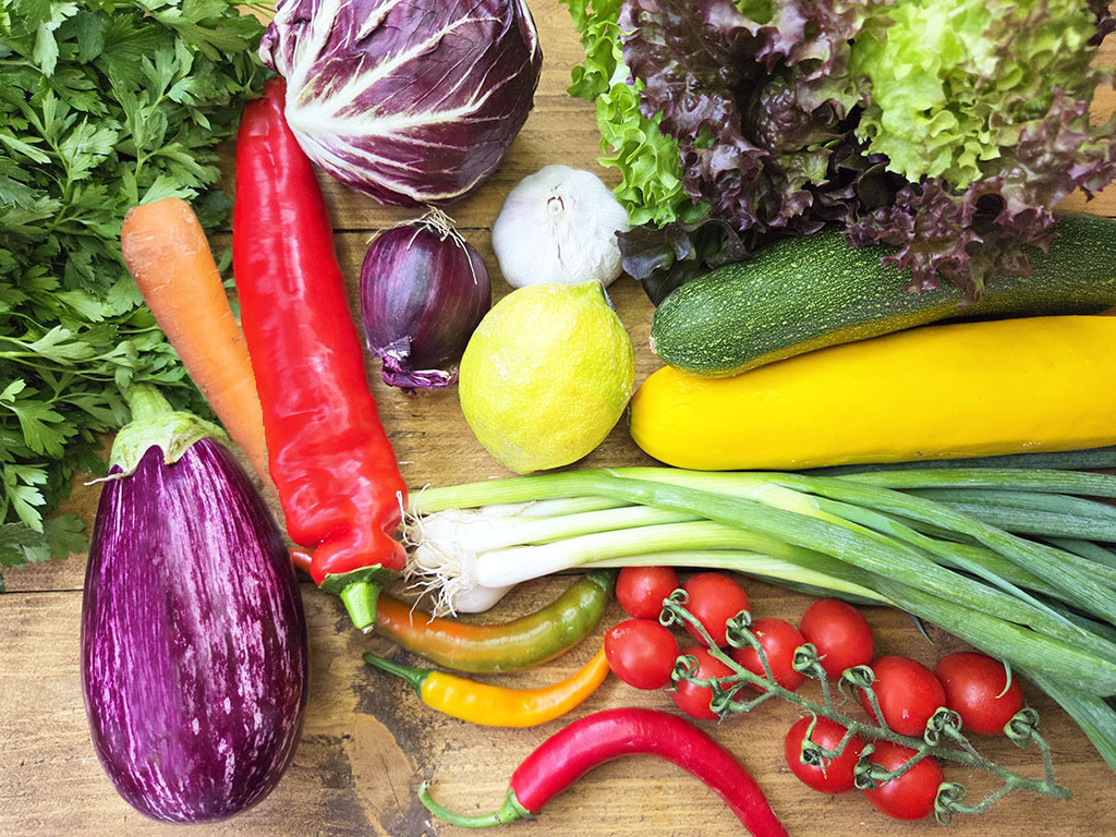 Assorted fresh vegetables on a wooden table: eggplants, red and yellow peppers, carrot, onion, garlic, lemon, zucchini, yellow squash, green onions, leafy greens, and cherry tomatoes.