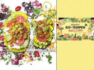 Two veggie tacos with lettuce, tomato, pickles, and crispy tempeh on a white wooden surface; on the right, a yellow panel with bio-tempeh packaging reading 'Bio-Tempeh Natur'