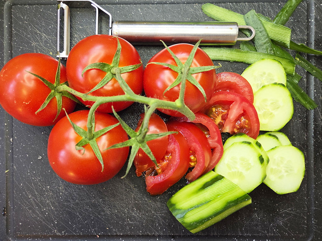 Tomatoes on the vine with cucumber slices and a peeler on a dark cutting board, ready for salad prep.